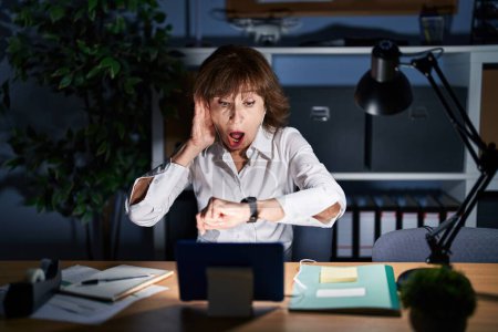 Téléchargez les photos : Femme d'âge moyen travaillant au bureau la nuit en regardant l'heure de la montre inquiet, peur de se retrouver en retard - en image libre de droit