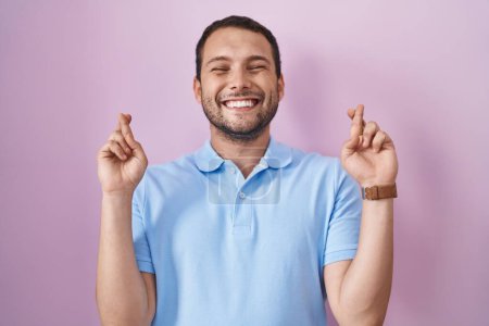 Téléchargez les photos : Homme hispanique debout sur fond rose geste doigt croisé souriant avec espoir et les yeux fermés. chance et concept superstitieux. - en image libre de droit