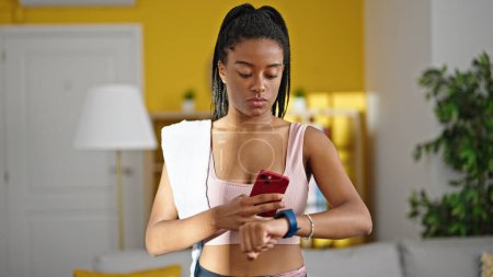 Téléchargez les photos : Femme afro-américaine portant des vêtements de sport à l'aide d'un smartphone regarder à la maison - en image libre de droit