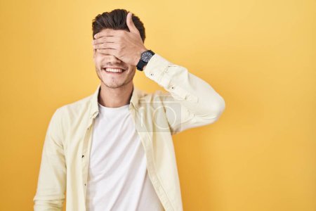 Téléchargez les photos : Jeune homme hispanique debout sur fond jaune souriant et riant avec la main sur le visage couvrant les yeux pour la surprise. concept aveugle. - en image libre de droit