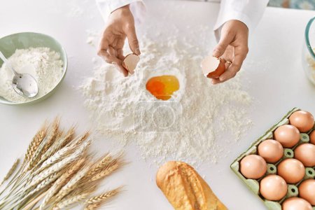Photo for Young woman wearing cook uniform cracking egg on flour at kitchen - Royalty Free Image