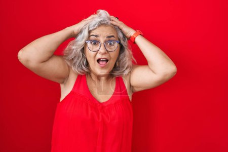 Photo for Middle age woman with grey hair standing over red background crazy and scared with hands on head, afraid and surprised of shock with open mouth - Royalty Free Image