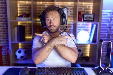 Téléchargez les photos : Homme d'âge moyen avec barbe jouant à des jeux vidéo portant un casque pointant des deux côtés avec les doigts, direction différente en désaccord - en image libre de droit