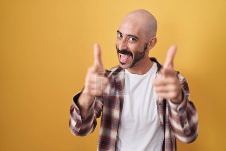 Téléchargez les photos : Homme hispanique avec barbe debout sur fond jaune pointant les doigts vers la caméra avec un visage heureux et drôle. bonne énergie et vibes. - en image libre de droit