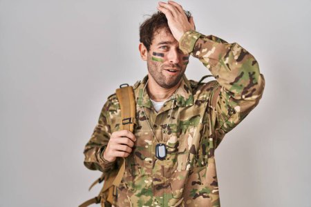 Téléchargez les photos : Jeune homme hispanique portant l'uniforme de l'armée de camouflage surpris avec la main sur la tête pour erreur, rappelez-vous erreur. oublié, mauvais concept de mémoire. - en image libre de droit