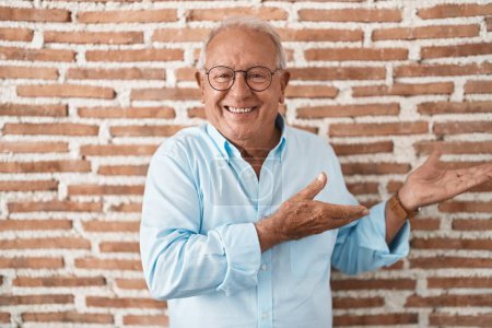 Téléchargez les photos : Homme âgé avec les cheveux gris debout sur le mur de briques invitant à entrer souriant naturel avec la main ouverte - en image libre de droit