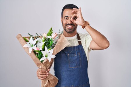 Foto de Hombre hispano con barba trabajando como florista haciendo buen gesto con la mano sonriente, ojos mirando a través de los dedos con cara feliz. - Imagen libre de derechos