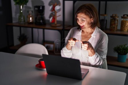 Téléchargez les photos : Femme hispanique du Moyen Age utilisant un ordinateur portable à la maison la nuit pointant les doigts vers la caméra avec un visage heureux et drôle. bonne énergie et vibes. - en image libre de droit