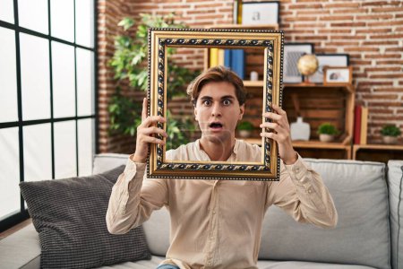 Photo for Young man holding empty frame afraid and shocked with surprise and amazed expression, fear and excited face. - Royalty Free Image
