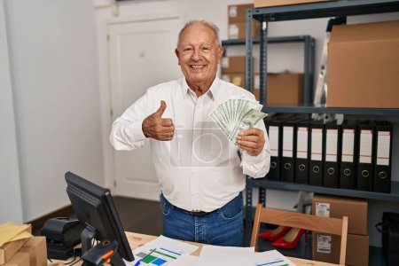 Foto de Hombre mayor con el pelo gris que trabaja en el comercio electrónico de la pequeña empresa sosteniendo dólares sonriendo feliz y positivo, pulgar hacia arriba haciendo excelente y signo de aprobación - Imagen libre de derechos