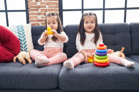 Foto de Adorable gemelas jugando con aros juego sosteniendo pato juguete sentado en el sofá en casa - Imagen libre de derechos