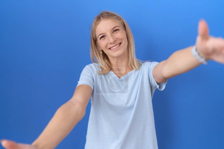 Téléchargez les photos : Jeune femme caucasienne portant un t-shirt bleu décontracté regardant la caméra souriant à bras ouverts pour un câlin. expression joyeuse embrassant le bonheur. - en image libre de droit