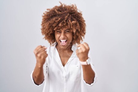 Foto de Mujer hispana joven con el pelo rizado de pie sobre fondo blanco enojado y loco levantando los puños frustrado y furioso mientras gritaba con ira. rabia y concepto agresivo. - Imagen libre de derechos