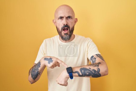 Foto de Hombre hispano con tatuajes de pie sobre fondo amarillo con prisa apuntando a ver el tiempo, la impaciencia, molesto y enojado por retraso de la fecha límite - Imagen libre de derechos