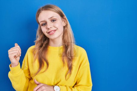 Téléchargez les photos : Jeune femme caucasienne debout sur fond bleu pointant vers le dos derrière avec la main et les pouces vers le haut, souriant confiant - en image libre de droit