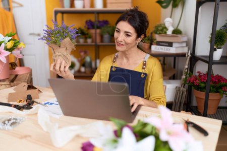 Téléchargez les photos : Moyen Age hispanique femme fleuriste en utilisant ordinateur portable tenant usine de lavande à la boutique de fleurs - en image libre de droit