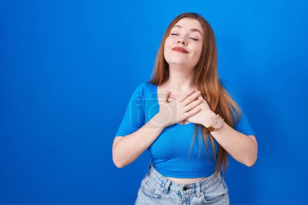 Photo for Redhead woman standing over blue background smiling with hands on chest with closed eyes and grateful gesture on face. health concept. - Royalty Free Image