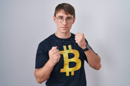 Photo for Caucasian blond man wearing bitcoin t shirt ready to fight with fist defense gesture, angry and upset face, afraid of problem - Royalty Free Image