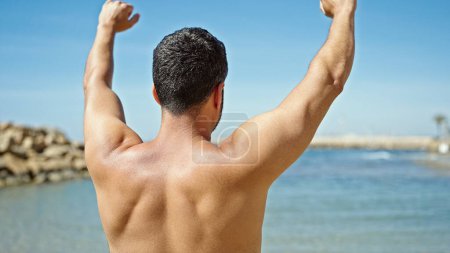 Foto de Joven turista hispano de pie sin camisa estirando los brazos en la playa - Imagen libre de derechos