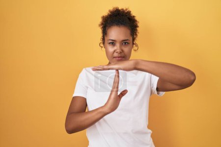 Photo for Young hispanic woman with curly hair standing over yellow background doing time out gesture with hands, frustrated and serious face - Royalty Free Image