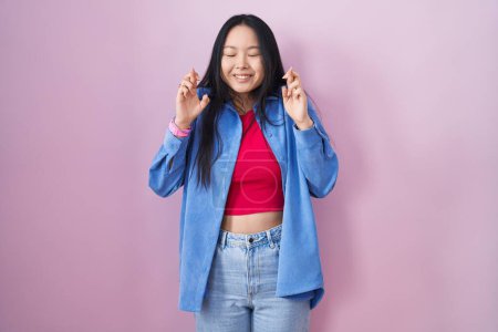 Téléchargez les photos : Jeune femme asiatique debout sur fond rose geste doigt croisé souriant avec espoir et les yeux fermés. chance et concept superstitieux. - en image libre de droit