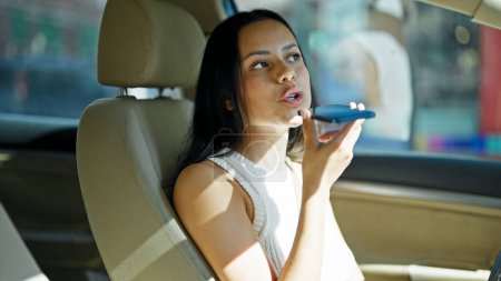 Photo for Young beautiful hispanic woman sending voice message by smartphone sitting on car at street - Royalty Free Image