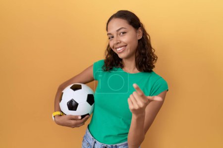 Foto de Joven mujer hispana sosteniendo la pelota haciendo señas ven aquí gesto con la mano invitando a la bienvenida feliz y sonriente - Imagen libre de derechos