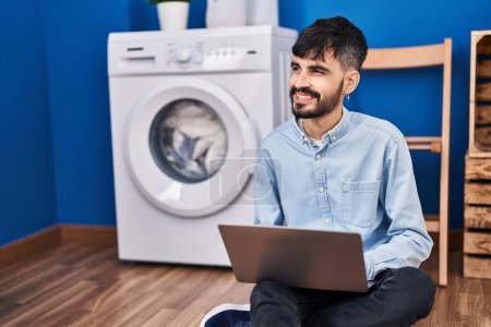 Téléchargez les photos : Jeune homme hispanique utilisant un ordinateur portable attendant la machine à laver à la buanderie - en image libre de droit