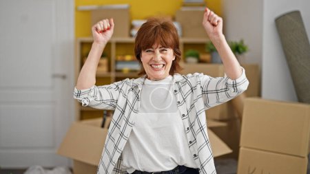 Téléchargez les photos : Femme d'âge moyen souriant confiance debout avec geste gagnant à la nouvelle maison - en image libre de droit