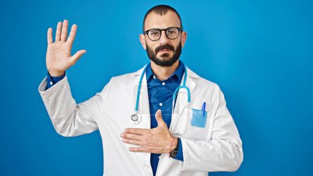 Téléchargez les photos : Jeune homme hispanique médecin faire un serment avec la main sur la poitrine sur fond bleu isolé - en image libre de droit