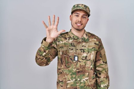 Téléchargez les photos : Jeune homme hispanique portant l'uniforme de camouflage de l'armée montrant et pointant vers le haut avec les doigts numéro cinq tout en souriant confiant et heureux. - en image libre de droit