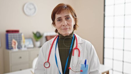Téléchargez les photos : Femme hispanique mature médecin debout avec une expression sérieuse à la clinique - en image libre de droit
