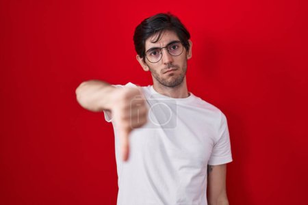 Photo for Young hispanic man standing over red background looking unhappy and angry showing rejection and negative with thumbs down gesture. bad expression. - Royalty Free Image