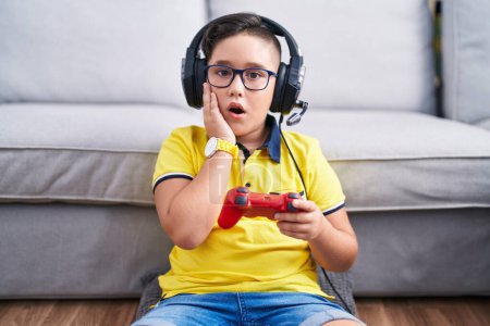 Téléchargez les photos : Jeune enfant hispanique jouant contrôleur de tenue de jeu vidéo portant casque peur et choqué, surprise et expression étonnante avec les mains sur le visage - en image libre de droit