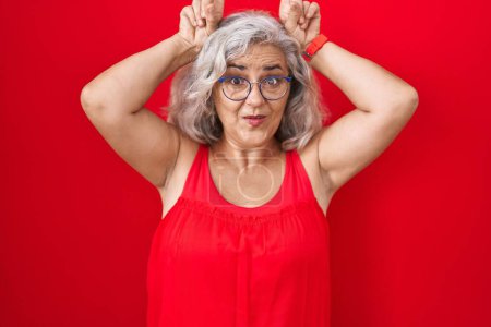 Téléchargez les photos : Femme d'âge moyen avec les cheveux gris debout sur fond rouge faisant un geste drôle avec le doigt sur la tête comme cornes de taureau - en image libre de droit