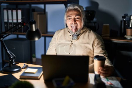 Téléchargez les photos : Homme d'âge moyen avec les cheveux gris qui travaillent au bureau la nuit en sortant la langue heureuse avec une expression drôle. concept d'émotion. - en image libre de droit