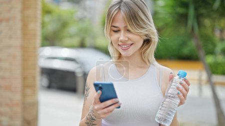 Téléchargez les photos : Jeune femme blonde utilisant un smartphone tenant une bouteille d'eau dans la rue - en image libre de droit