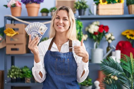 Foto de Mujer rubia joven que trabaja en floristería sosteniendo dólares sonriendo feliz y positivo, pulgar hacia arriba haciendo excelente y signo de aprobación - Imagen libre de derechos