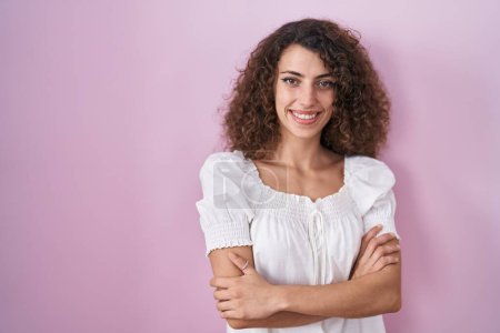 Téléchargez les photos : Femme hispanique avec les cheveux bouclés debout sur fond rose visage heureux souriant avec les bras croisés regardant la caméra. personne positive. - en image libre de droit