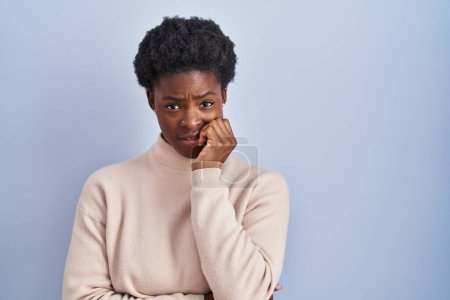 Foto de African american woman standing over blue background looking stressed and nervous with hands on mouth biting nails. anxiety problem. - Imagen libre de derechos