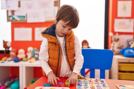Foto de Adorable chico caucásico jugando con matemáticas juego de puzzle sentado en la mesa en el jardín de infantes - Imagen libre de derechos