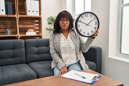 Foto de Mujer hispana que trabaja en la oficina de terapia con actitud de pensar en el reloj y expresión sobria que parece segura de sí misma - Imagen libre de derechos
