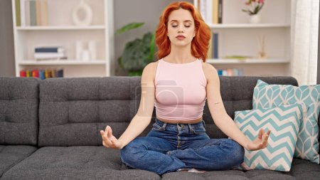 Foto de Mujer pelirroja joven haciendo ejercicio de yoga sentado en el sofá en casa - Imagen libre de derechos