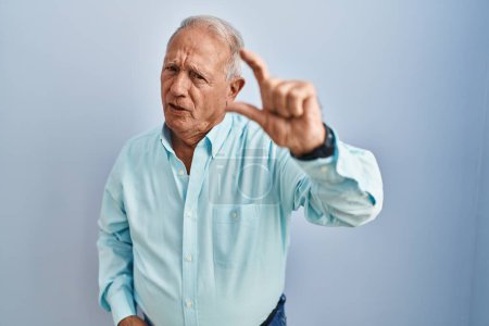 Téléchargez les photos : Homme âgé avec les cheveux gris debout sur fond bleu geste souriant et confiant avec la main faisant signe de petite taille avec les doigts à la recherche et la caméra. concept de mesure. - en image libre de droit