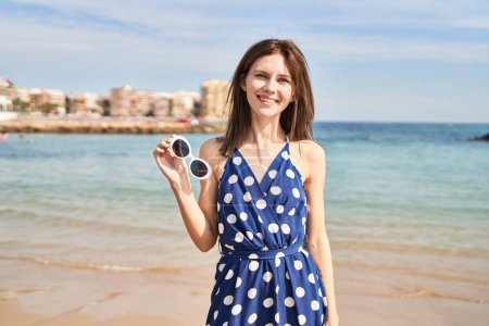 Téléchargez les photos : Jeune femme blonde touriste souriant confiant tenant des lunettes de soleil à la plage - en image libre de droit