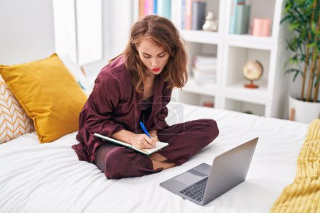 Photo for Young beautiful hispanic woman using laptop writing on notebook at bedroom - Royalty Free Image