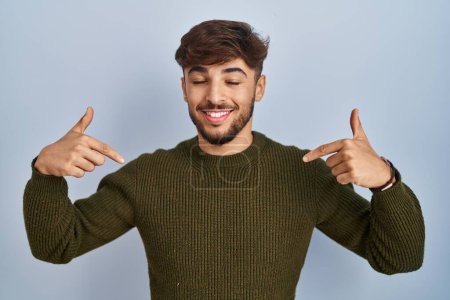 Foto de Hombre árabe con barba de pie sobre fondo azul mirando confiado con sonrisa en la cara, señalándose con los dedos orgullosos y felices. - Imagen libre de derechos