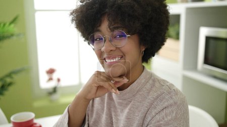 Téléchargez les photos : Jeune femme afro-américaine souriante confiante assise sur la table à la maison - en image libre de droit