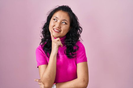 Foto de Mujer asiática joven de pie sobre fondo rosa con la mano en la barbilla pensando en la pregunta, la expresión pensativa. Cara sonriente y pensativa. concepto de duda. - Imagen libre de derechos