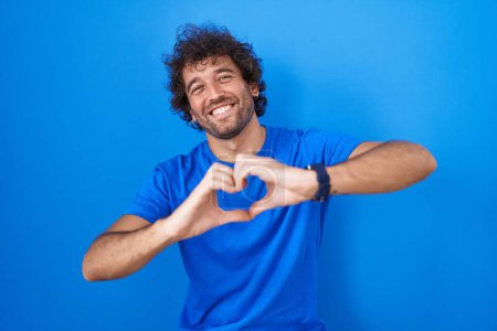 Téléchargez les photos : Jeune homme hispanique debout sur fond bleu souriant dans l'amour faisant forme de symbole de coeur avec les mains. concept romantique. - en image libre de droit